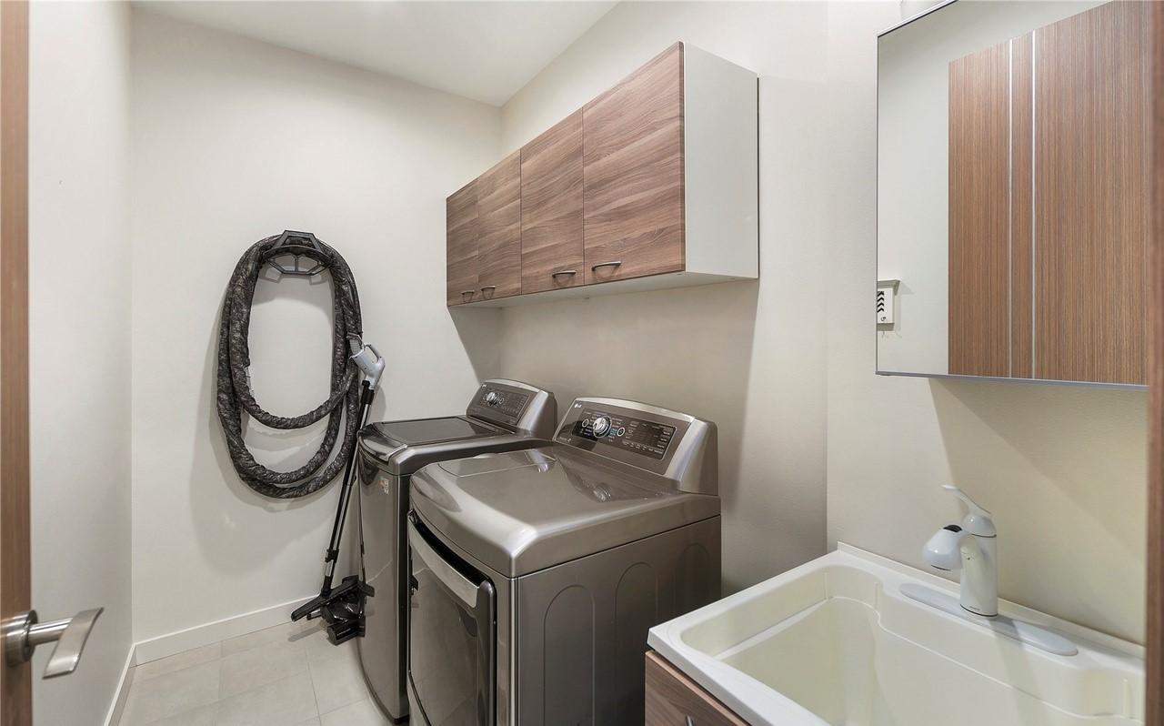 Laundry room with stacked washer and dryer, utility sink, overhead cabinets, and a coiled hose on a wall mount.