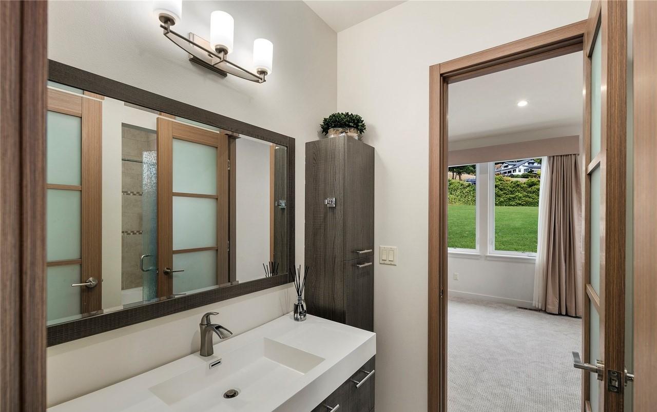 Modern bathroom with double vanity, frosted glass doors, and view into bedroom with large windows.
