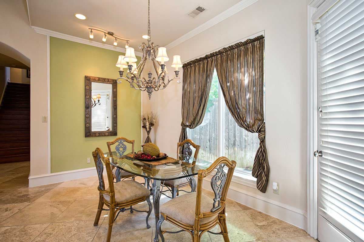 Dining area with glass table, four chairs, chandelier, and window with striped drapes. Stairs visible in background.