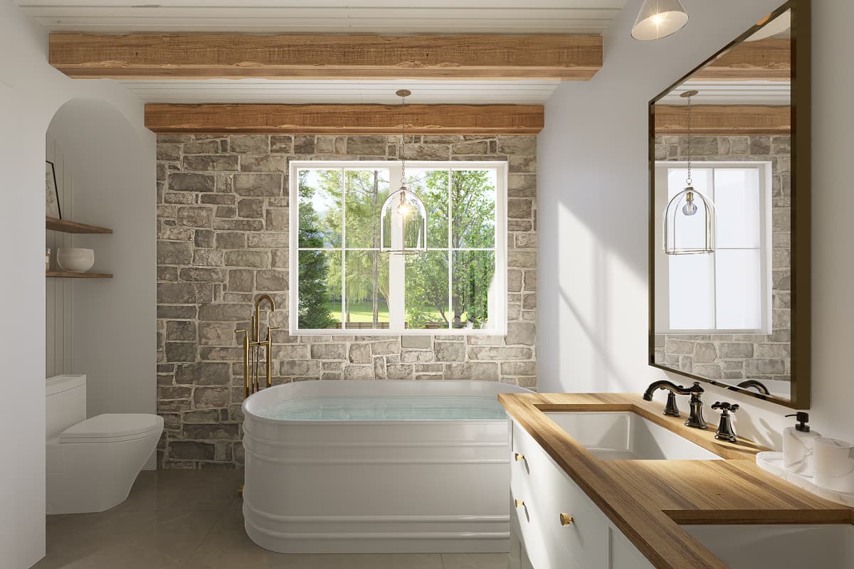 A rustic bathroom with a white tub in front of a stone wall. A window offers a view of green foliage, and wooden beams adorn the ceiling.