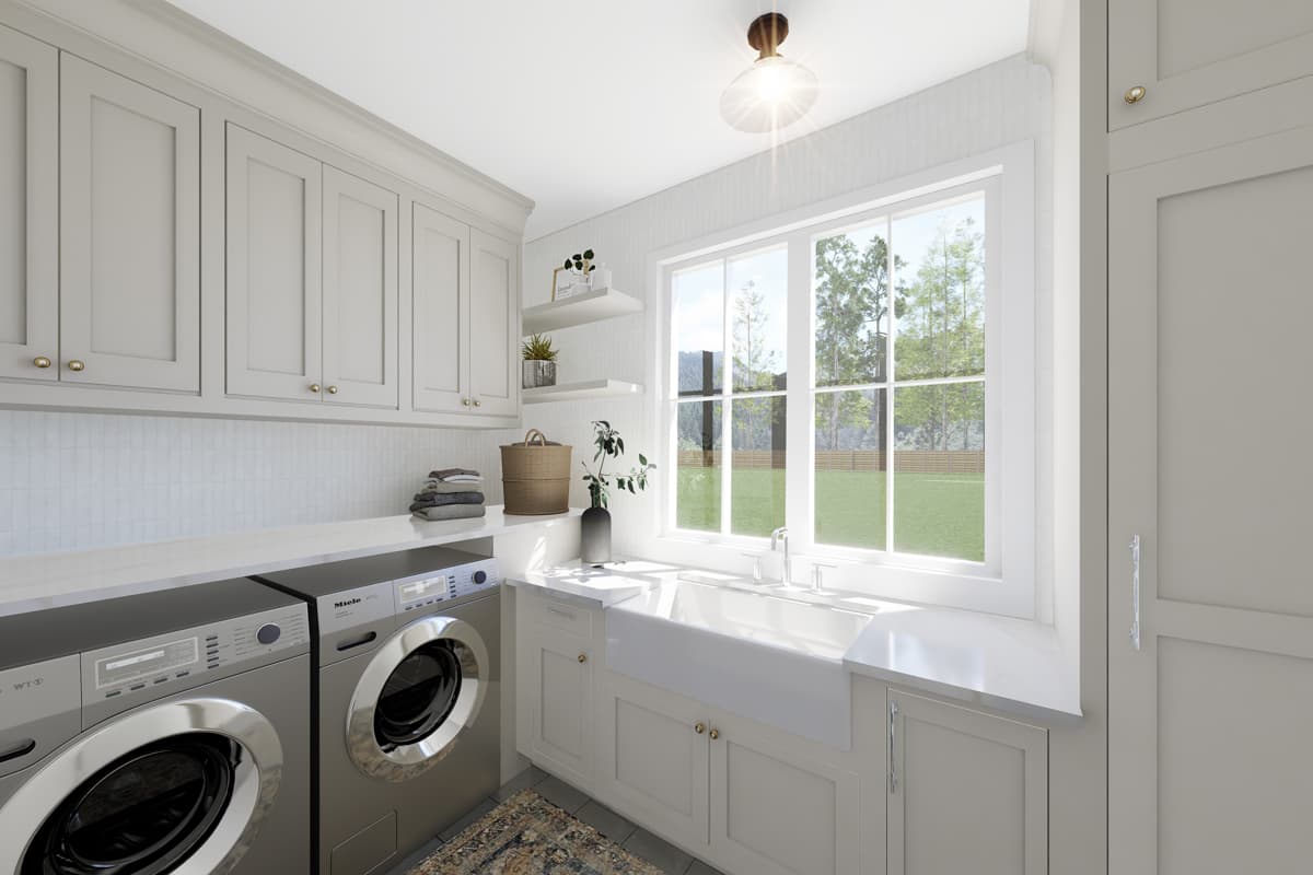 A modern, bright laundry room featuring a washer and dryer, a farmhouse sink, and large windows overlooking greenery. Upper cabinets and shelves are visible.