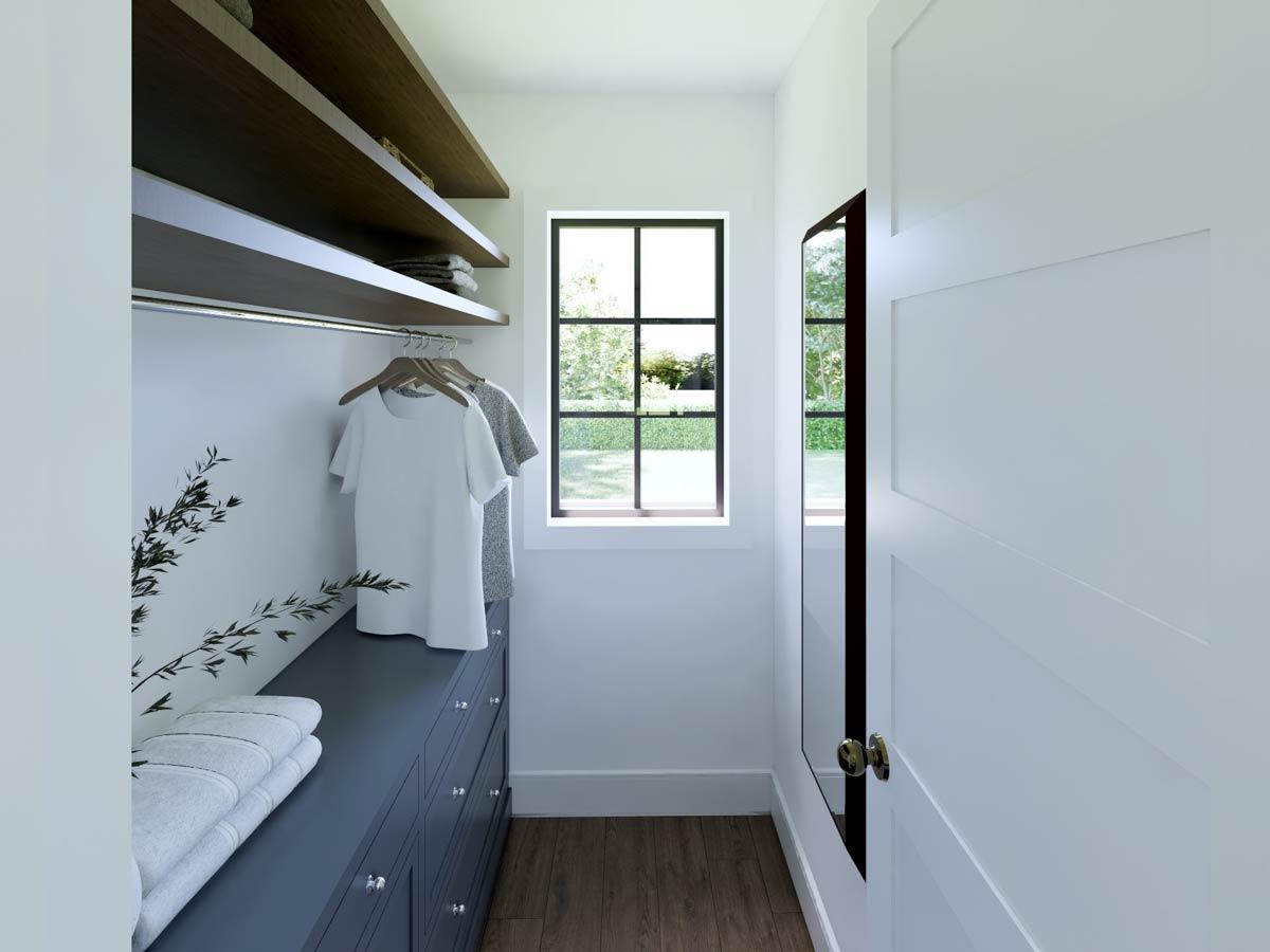 A walk-in closet with shelves, a drawer unit, and a rod with clothes. A window provides a view of a green yard and tree line.