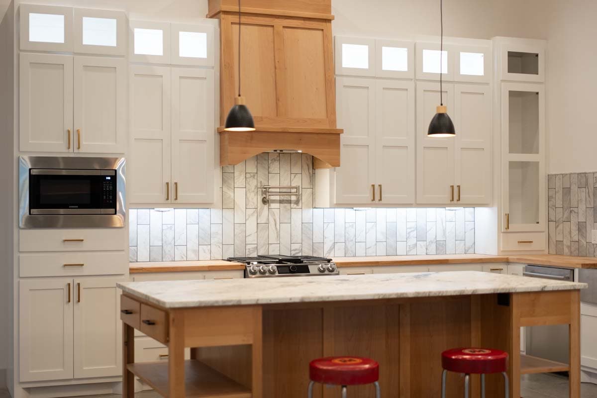Kitchen with white shaker cabinets, wood range hood, marble tile backsplash, gas range, and island with marble countertop.