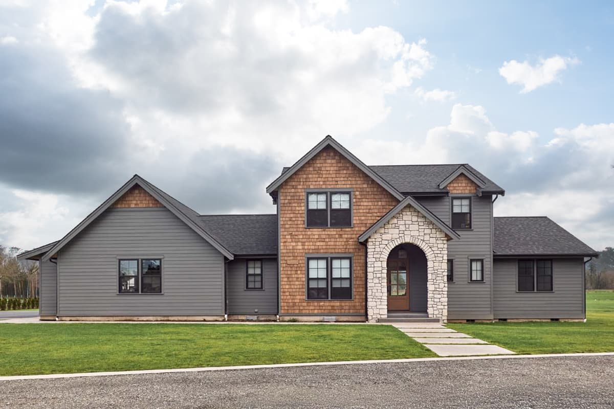 House plan exterior. Two-story home with a gabled roof, stone arch entryway, and cedar shake siding. Gray siding and dark windows.