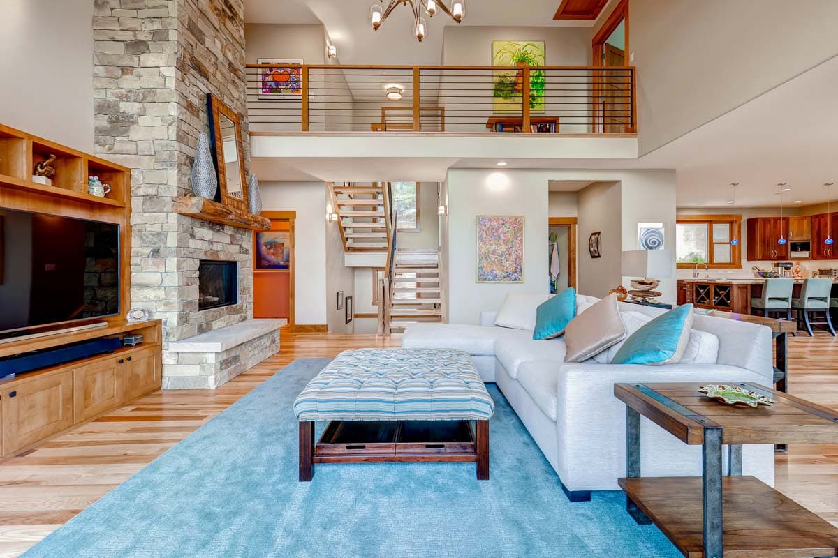 Interior view of living area with stone fireplace, wood shelving, open staircase, loft, and kitchen.