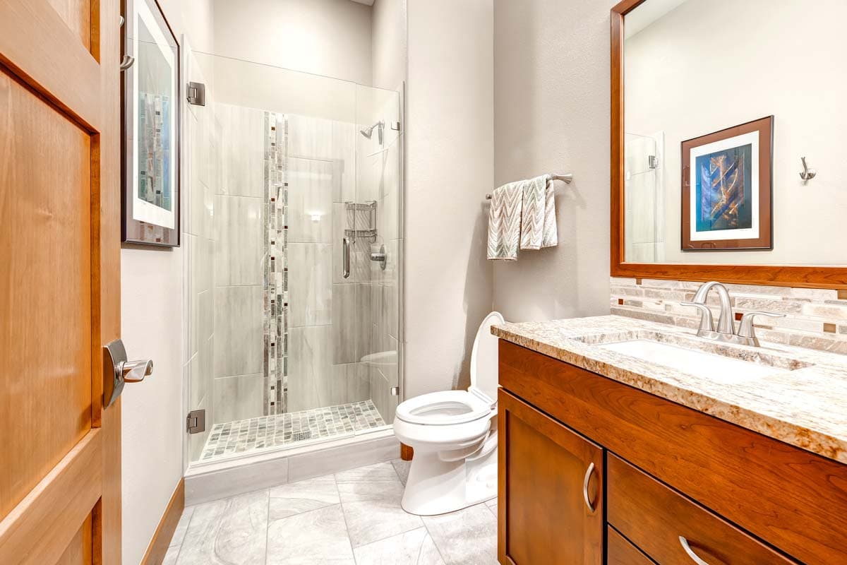Modern bathroom interior with glass-enclosed shower, wooden vanity with granite countertop, and tiled flooring.