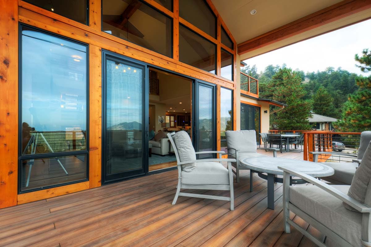 Modern house plan exterior with large windows, wooden beams, deck, and balcony overlooking a wooded landscape.