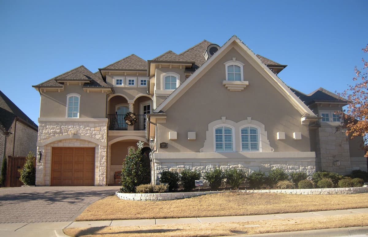 House plan exterior: Two-story home with stucco and stone facade, arched garage door, and balcony with ornate railing.