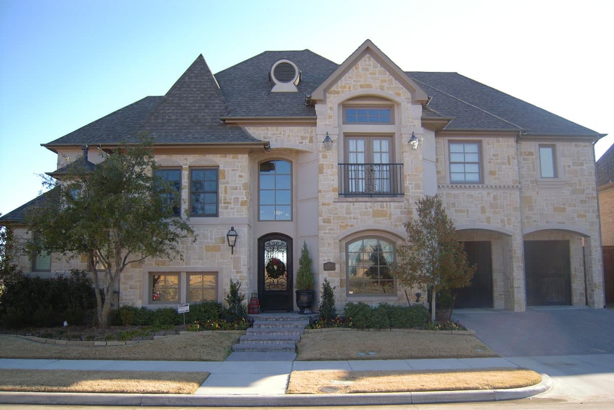 House plan exterior: two-story stone home with prominent gables, arched windows, second-floor balcony, and a three-car garage.