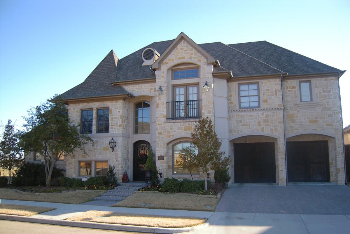 House plan exterior: two-story stone residence with a hipped roof, arched entryway, balcony, and a two-car side-entry garage.