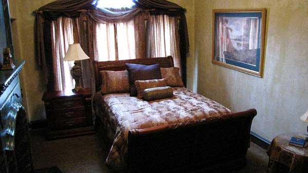 Bedroom interior with sleigh bed, patterned bedding, bedside table with lamp, and decorative curtain treatments.