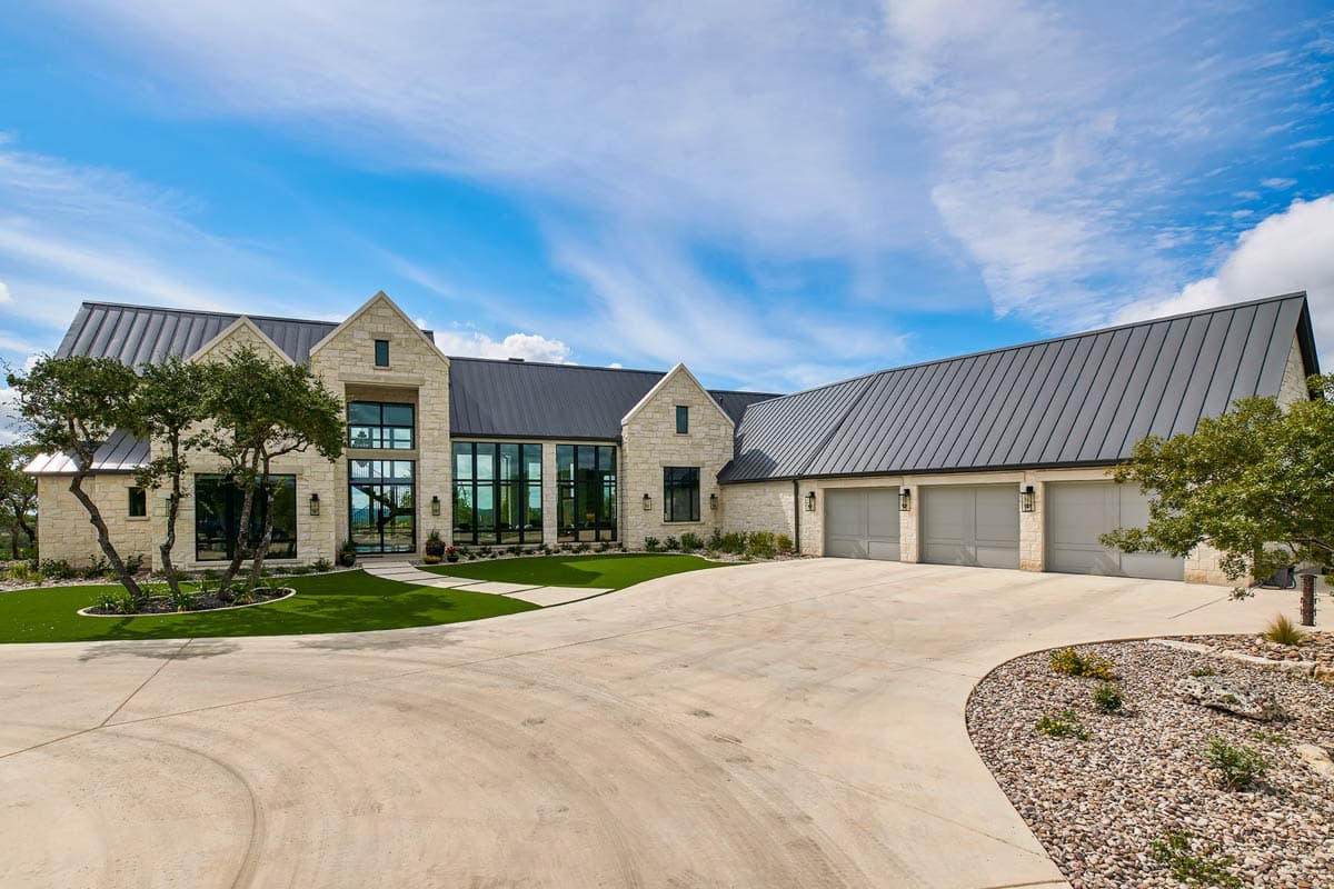 House plan exterior. Modern style, single story, with gable roof, large windows, and a three-car garage. Stone facade with metal roof.