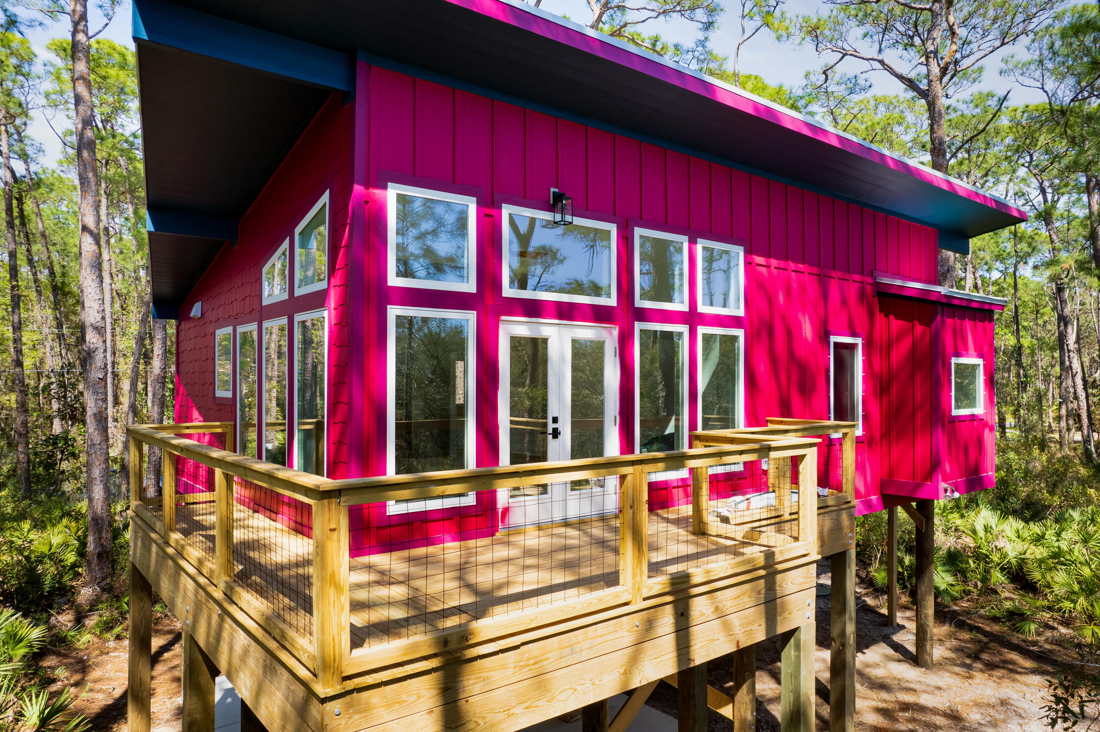 Modern house plan exterior with magenta siding, large windows, double doors, and a wooden deck on stilts.