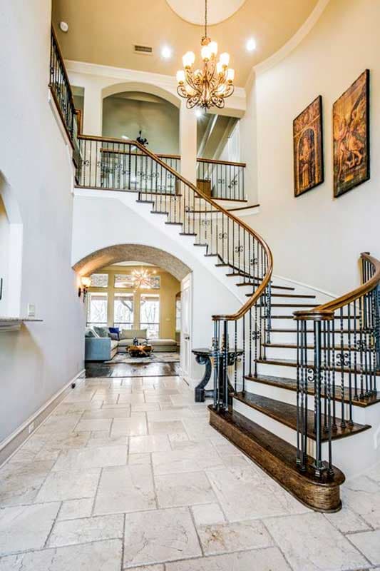 Interior view of a grand foyer with a curved staircase, wrought iron railings, tiled floor, and a chandelier.