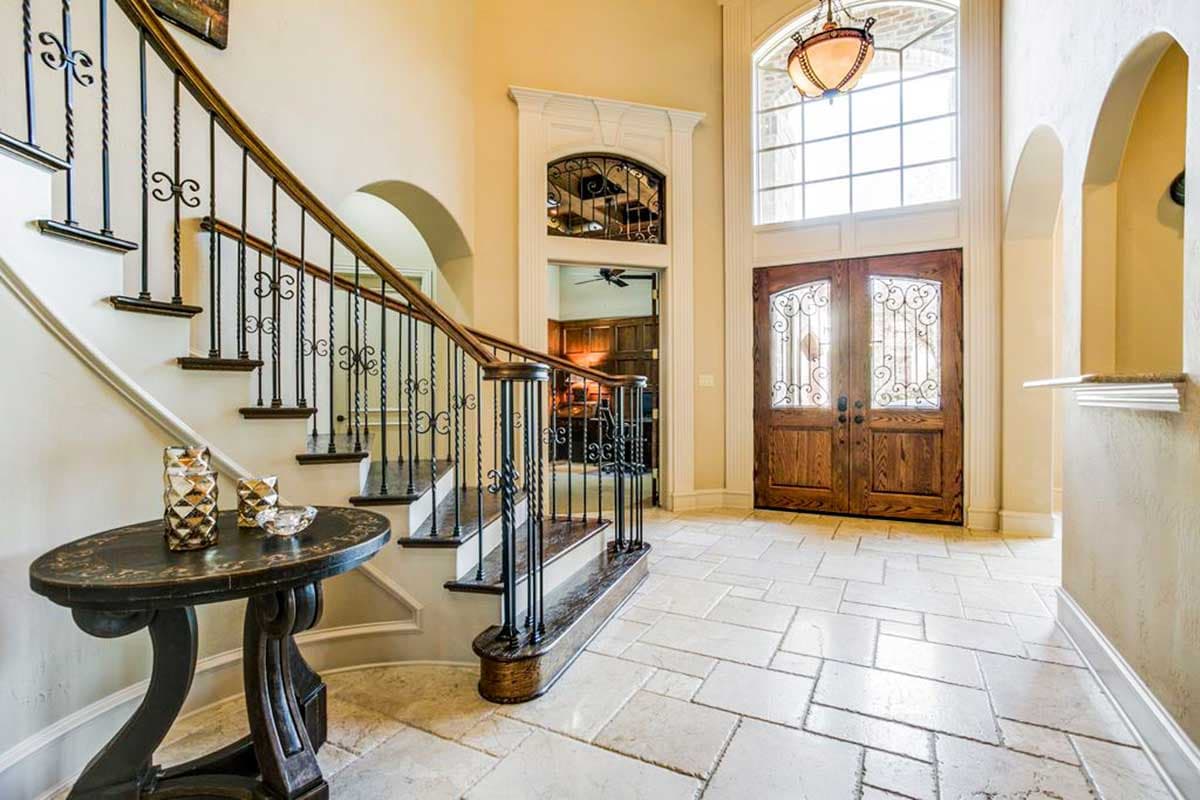 Interior view of a grand foyer with a sweeping staircase, wrought iron railing, and double wood doors beneath a large Palladian window.