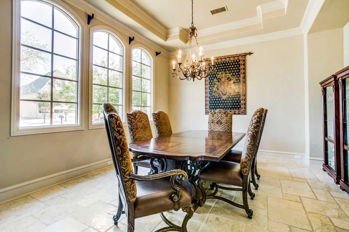 Dining room with large arched windows, ornate chandelier, tapestry, and dark wood table with upholstered chairs.