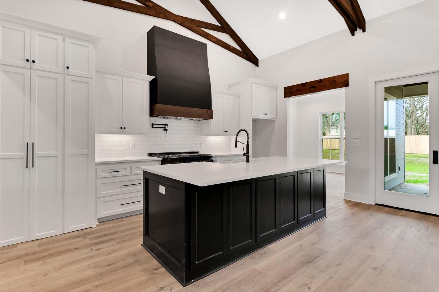 Kitchen with vaulted ceiling, exposed beams, island, and black range hood. White cabinetry and subway tile backsplash.