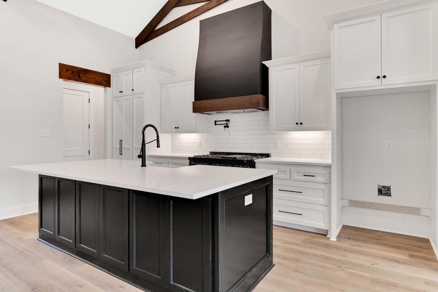 Kitchen with vaulted ceiling, exposed beams, large island, black range hood, and white cabinetry.