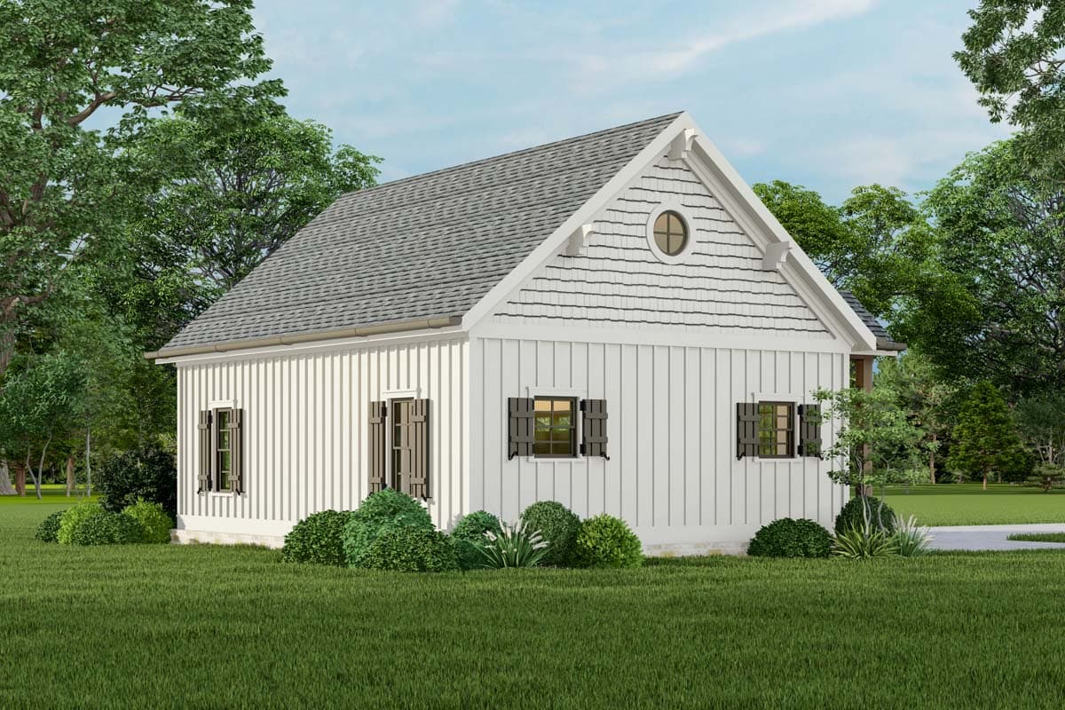 A white clapboard cottage with a gray roof and black shutters sits among green trees and grass, featuring a circular attic window.