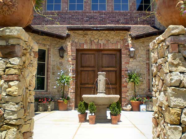 House plan exterior with stone pillars, a fountain, and double wooden doors. Brick facade with multi-pane windows above.
