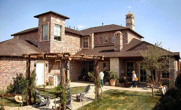 House plan exterior showing a two-story brick home with a prominent tower, multiple gables, and a rustic pergola over a patio.