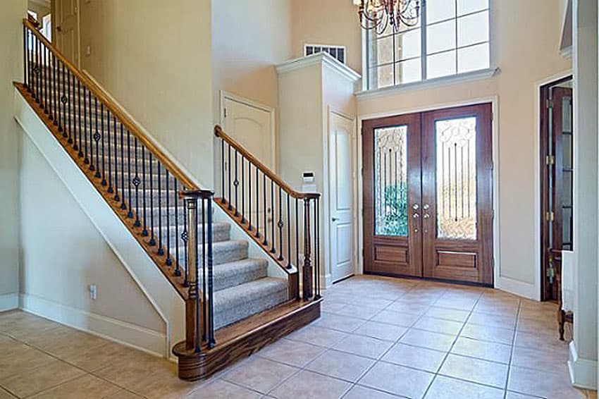 Interior view of a home foyer with a carpeted staircase, ornate metal balusters, and double wooden entry doors with glass panels.