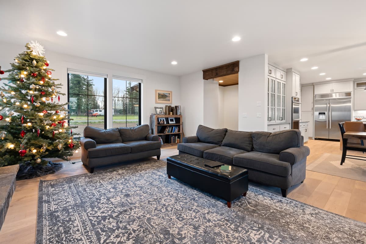 Living area with sectional sofa, ottoman, bookshelf, and Christmas tree, adjacent to kitchen with stainless steel appliances and island.