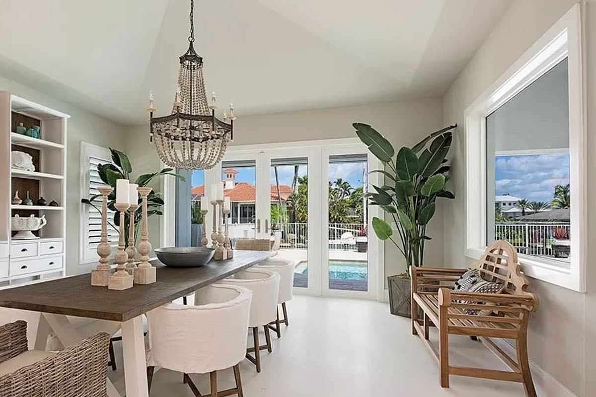 Dining area with vaulted ceiling, chandelier, long table, white chairs, and French doors opening to a pool.