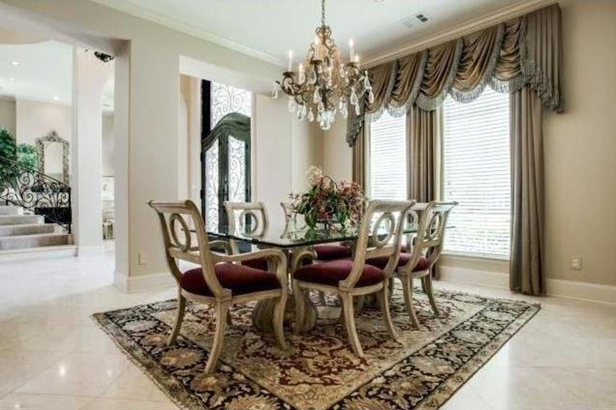 Dining room with glass-top table, six chairs, ornate chandelier, draped window treatments, and ornate rug. Staircase visible.