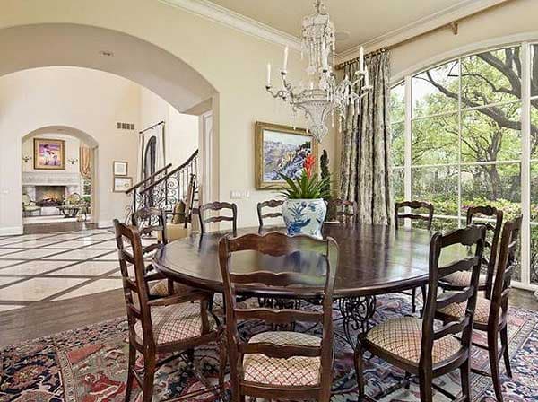 Dining room with oval table, upholstered chairs, chandelier, arched entryway to living room with fireplace, and large arched window.