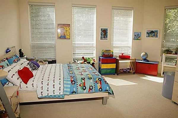 Interior view of a child's bedroom with a twin bed, dresser, toy storage, and multiple windows with blinds.