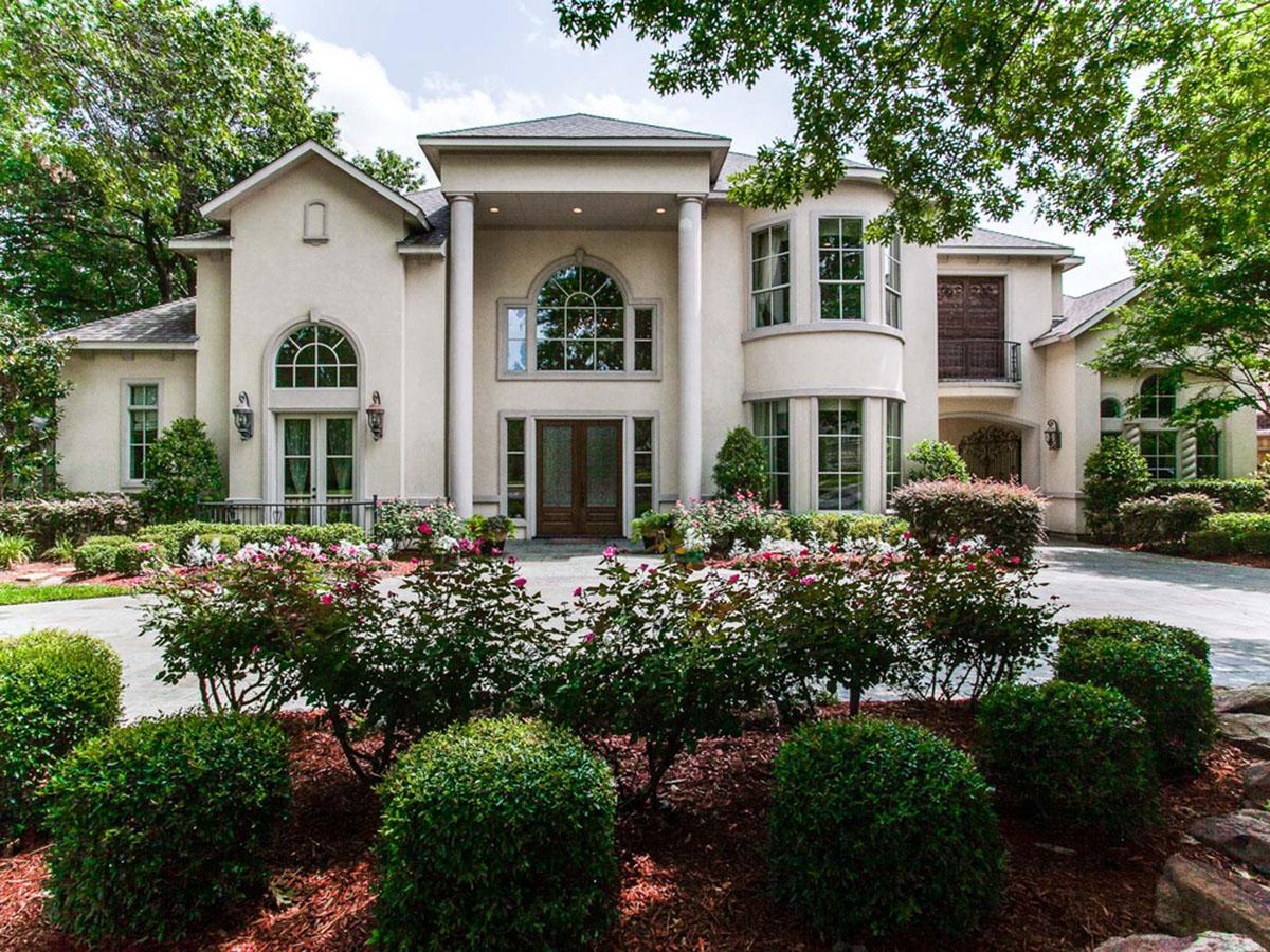 House plan exterior. Two-story, stucco facade with large arched windows, multiple columns, and a covered entry. Landscaping features flowering bushes.