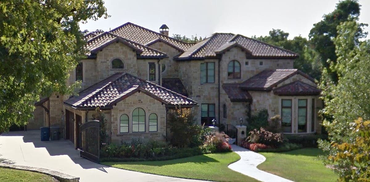 House plan exterior. Two-story Mediterranean-style home with multiple rooflines, arched windows, and a gated entrance with a long driveway.