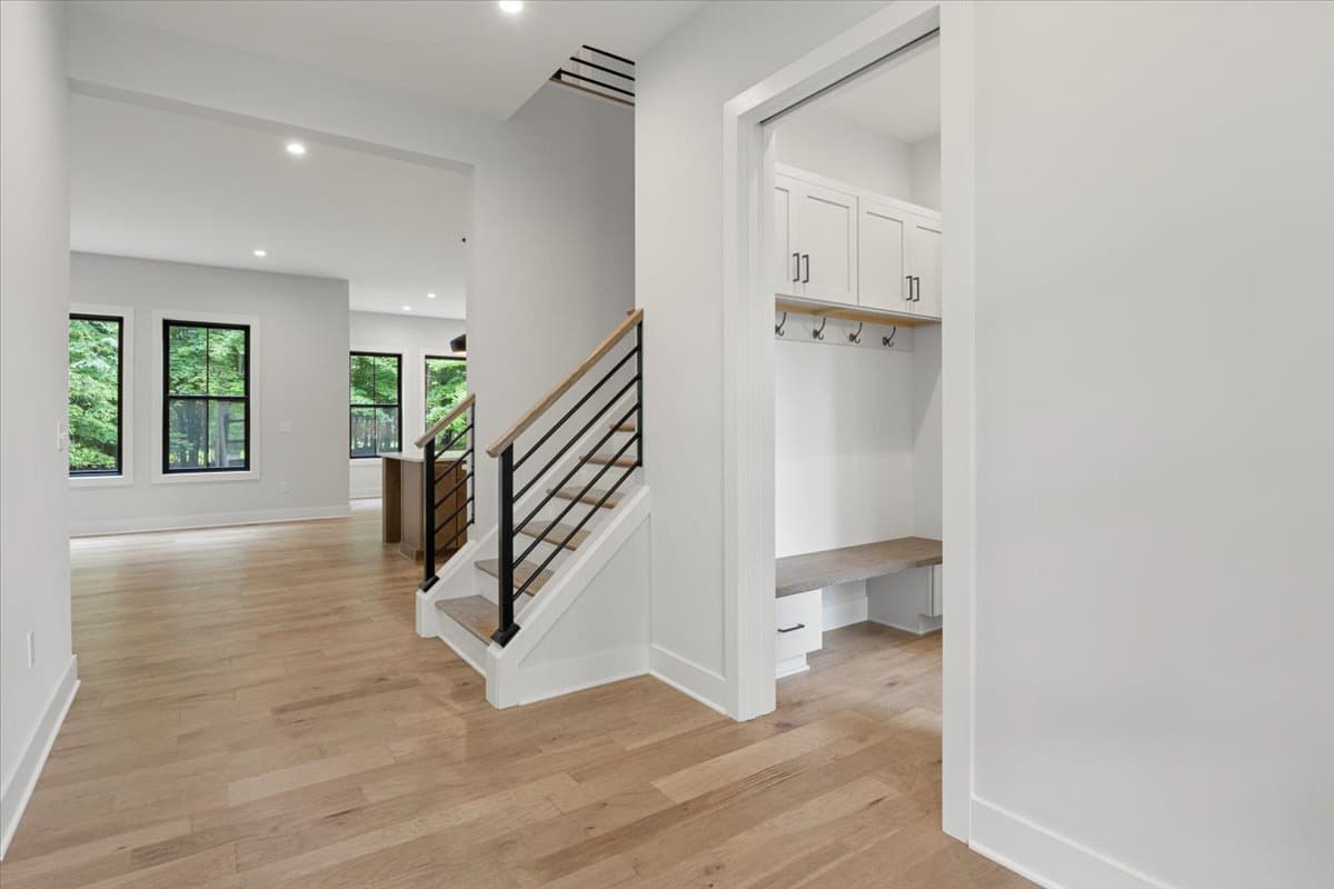Interior view showing staircase with black railing, mudroom with bench and cabinets, and open living area with windows.