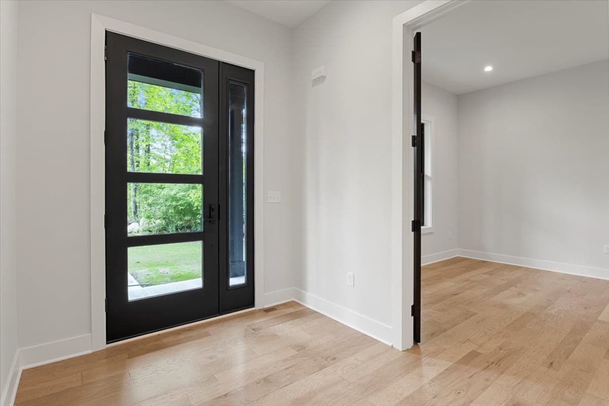 Modern entryway with black double doors featuring glass panels, wood flooring, and view into an adjacent room.