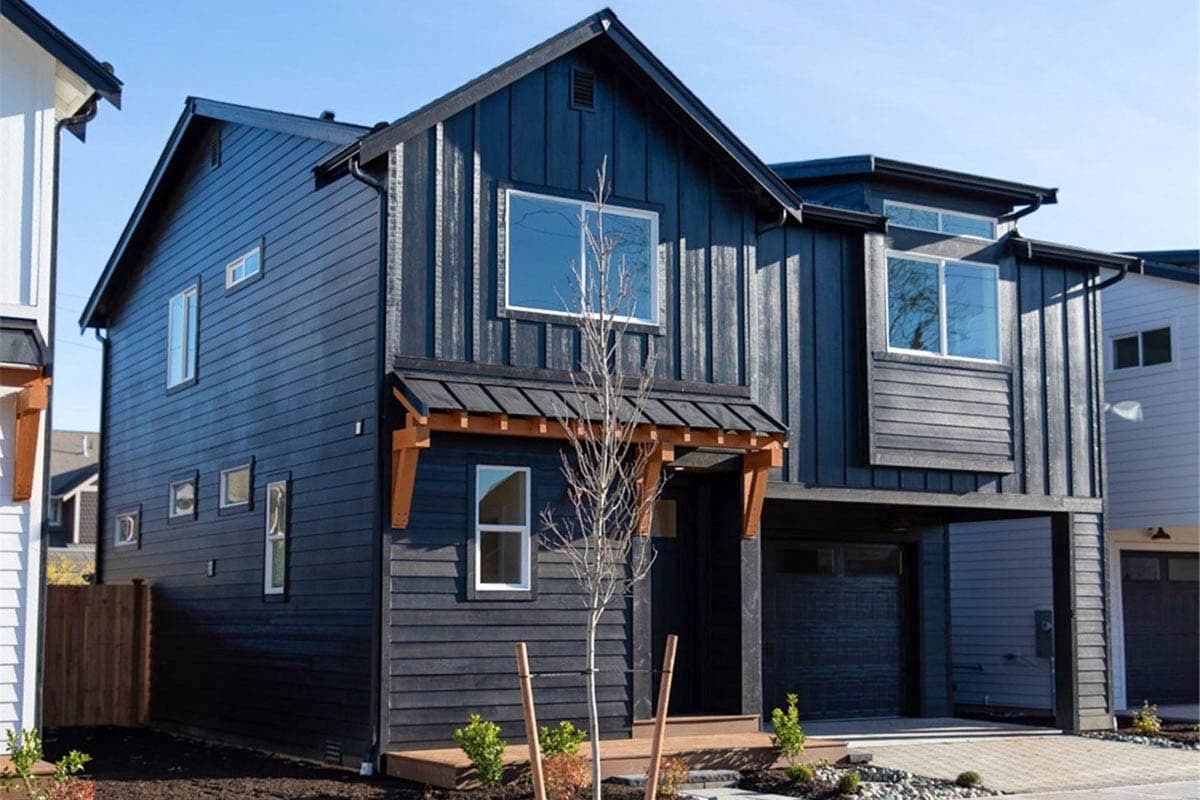 House plan exterior. Two-story modern house with dark siding, a gabled roof, and a covered porch. Attached garage has a flat roof.