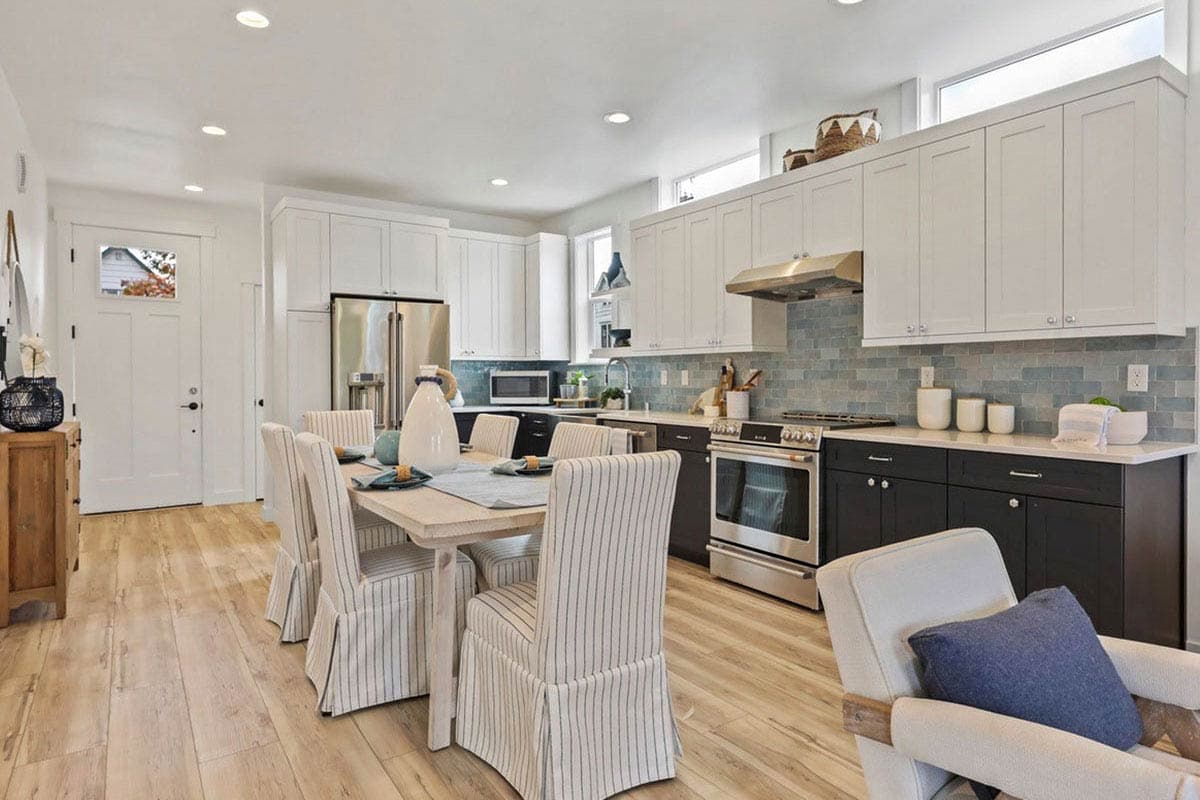 Open-concept kitchen and dining area with white cabinets, dark base cabinets, stainless steel appliances, and a dining table with slipcovered chairs.