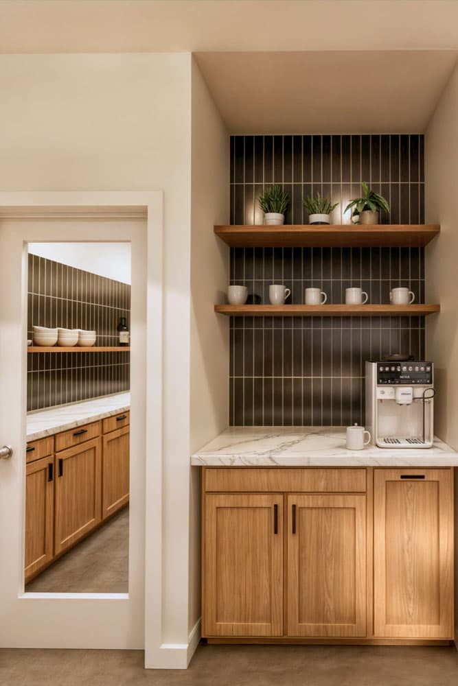 A coffee station with a built-in espresso machine, wooden shelves, and cabinets. A doorway reveals a pantry with similar cabinetry.