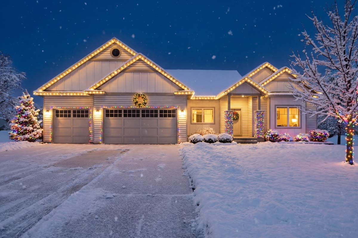 A snow-covered house at night, decorated with Christmas lights and wreaths. A decorated tree and bushes add to the festive scene.