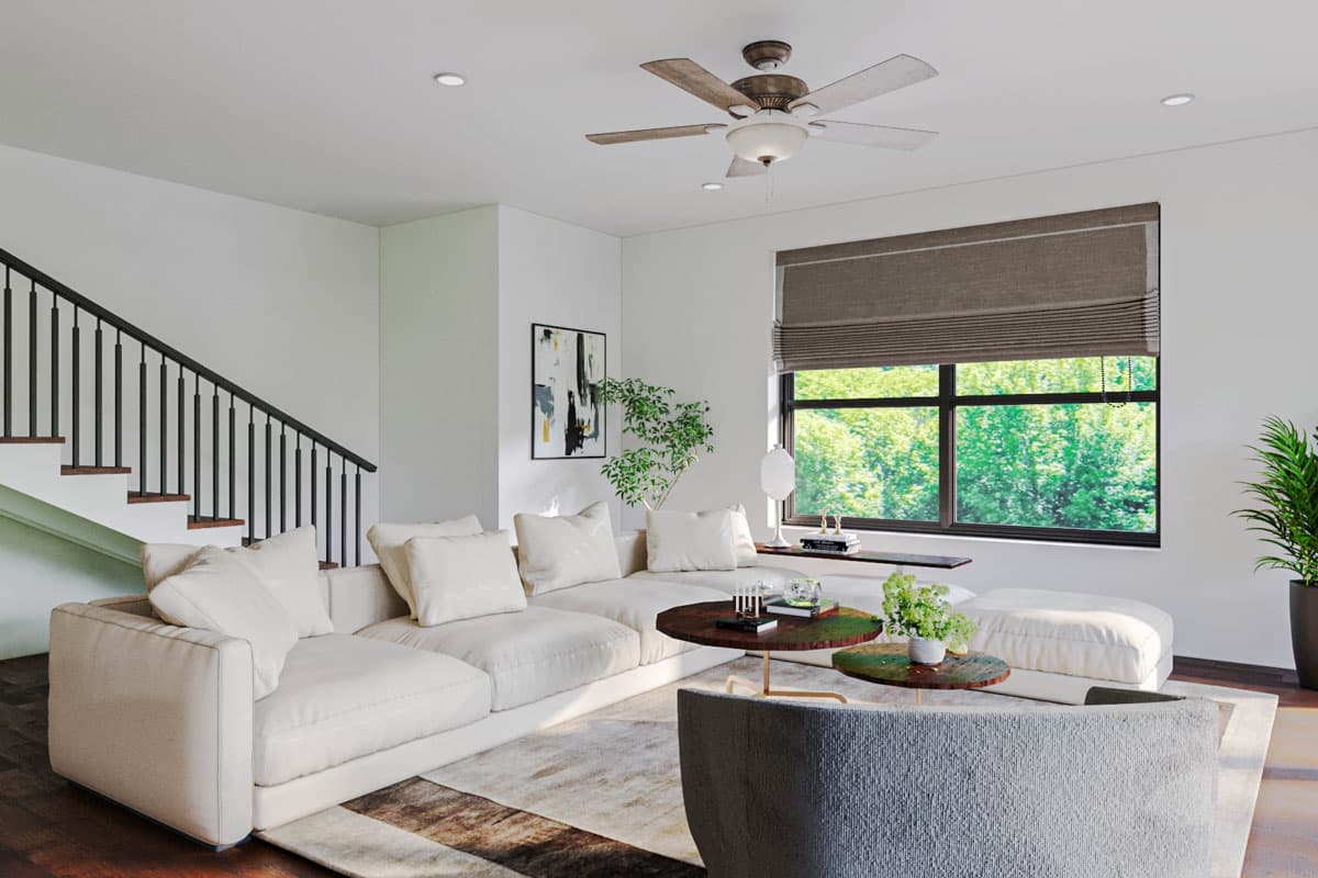 Interior view of a living room with a sectional sofa, staircase with black railing, ceiling fan, and large window with natural light.