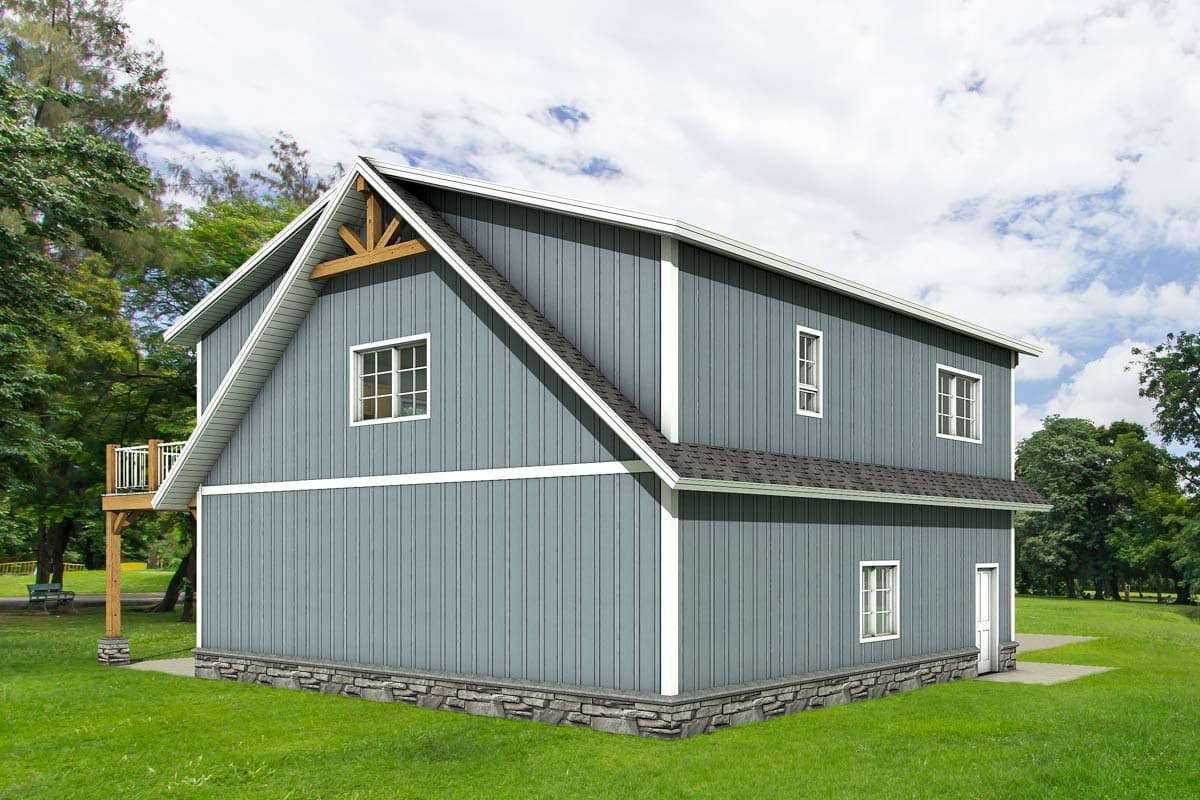 A two-story gray barn-style building with white trim, a stone base, and a wooden balcony is set on a grassy lawn under a cloudy sky.