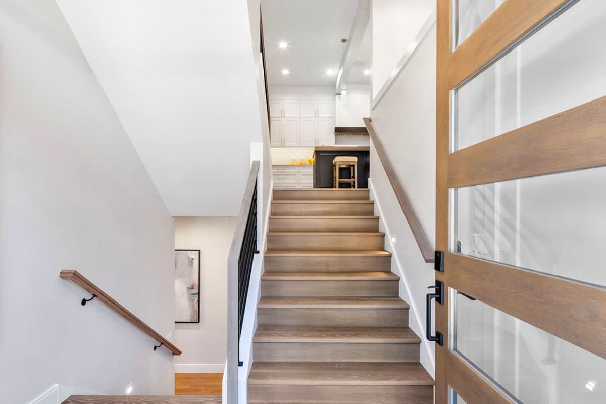Modern staircase with wood treads, glass and wood door, leading to a kitchen with white cabinets and island.