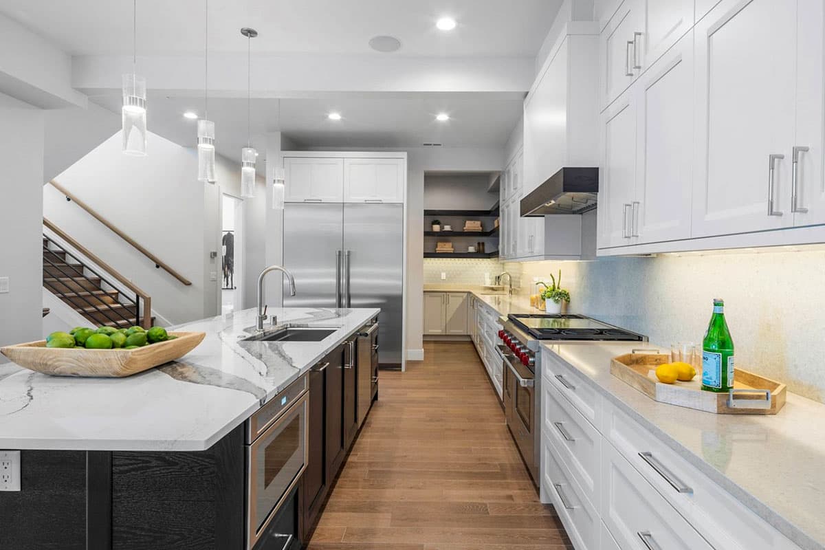 Modern kitchen with island, stainless steel appliances, white cabinetry, and wood flooring. Staircase visible.