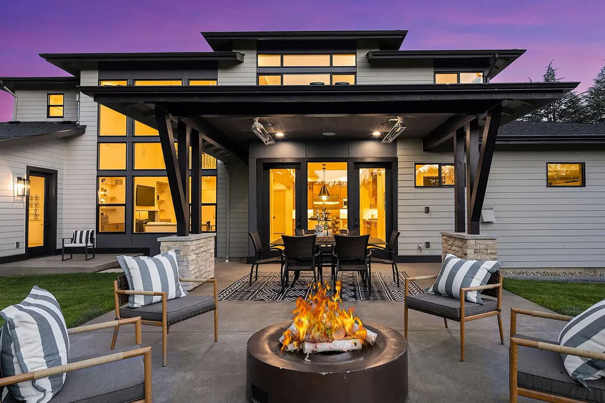 Modern house exterior with two stories, large windows, covered patio with outdoor dining, and a fire pit.