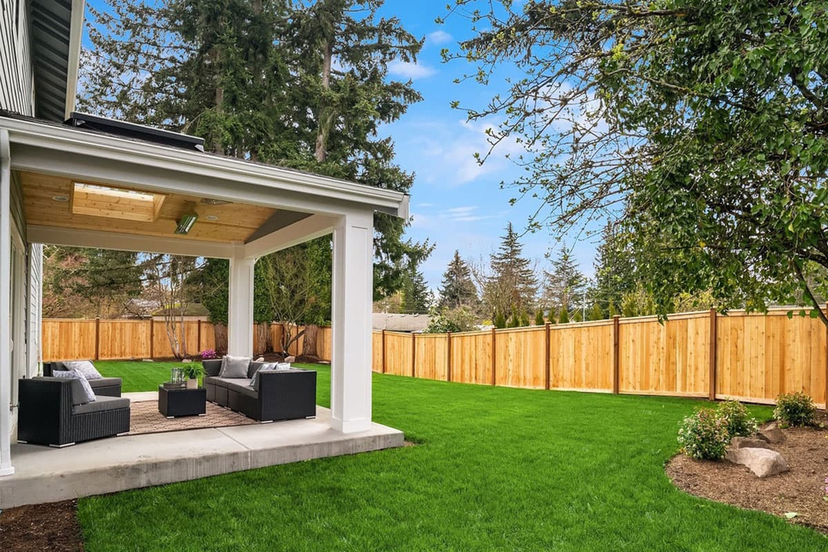Covered patio with seating area and wood-paneled ceiling overlooks a fenced backyard with green lawn and mature trees.