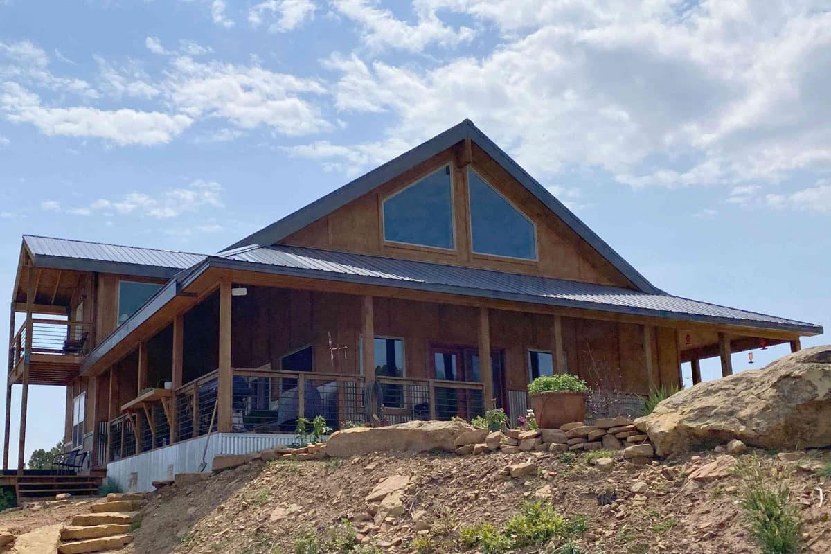 Modern house plan exterior with prominent gable, metal roof, wrap-around porch, and upper-level balcony.