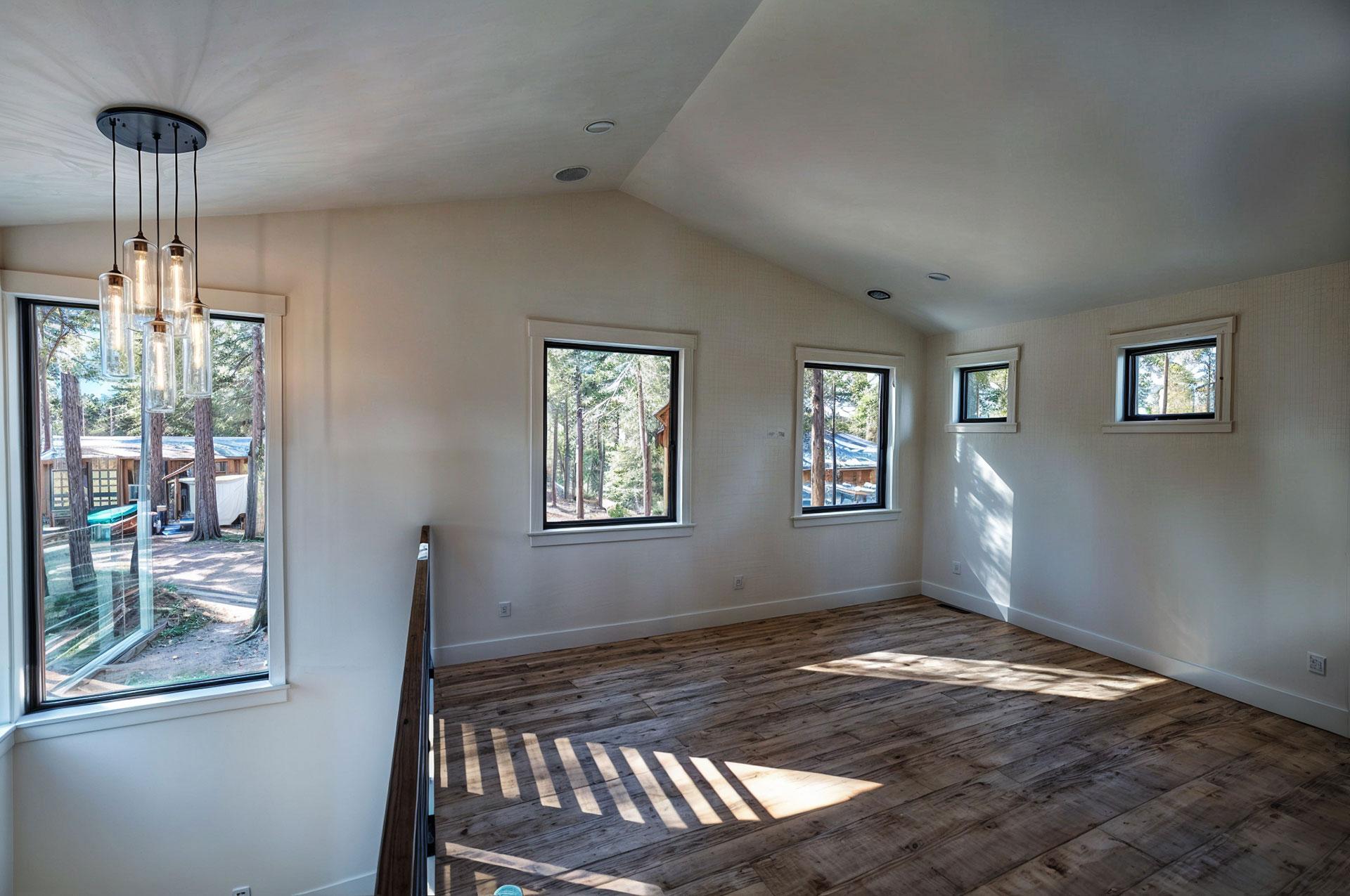 Interior view of a room with vaulted ceiling, wood floors, and multiple windows showcasing a wooded exterior.