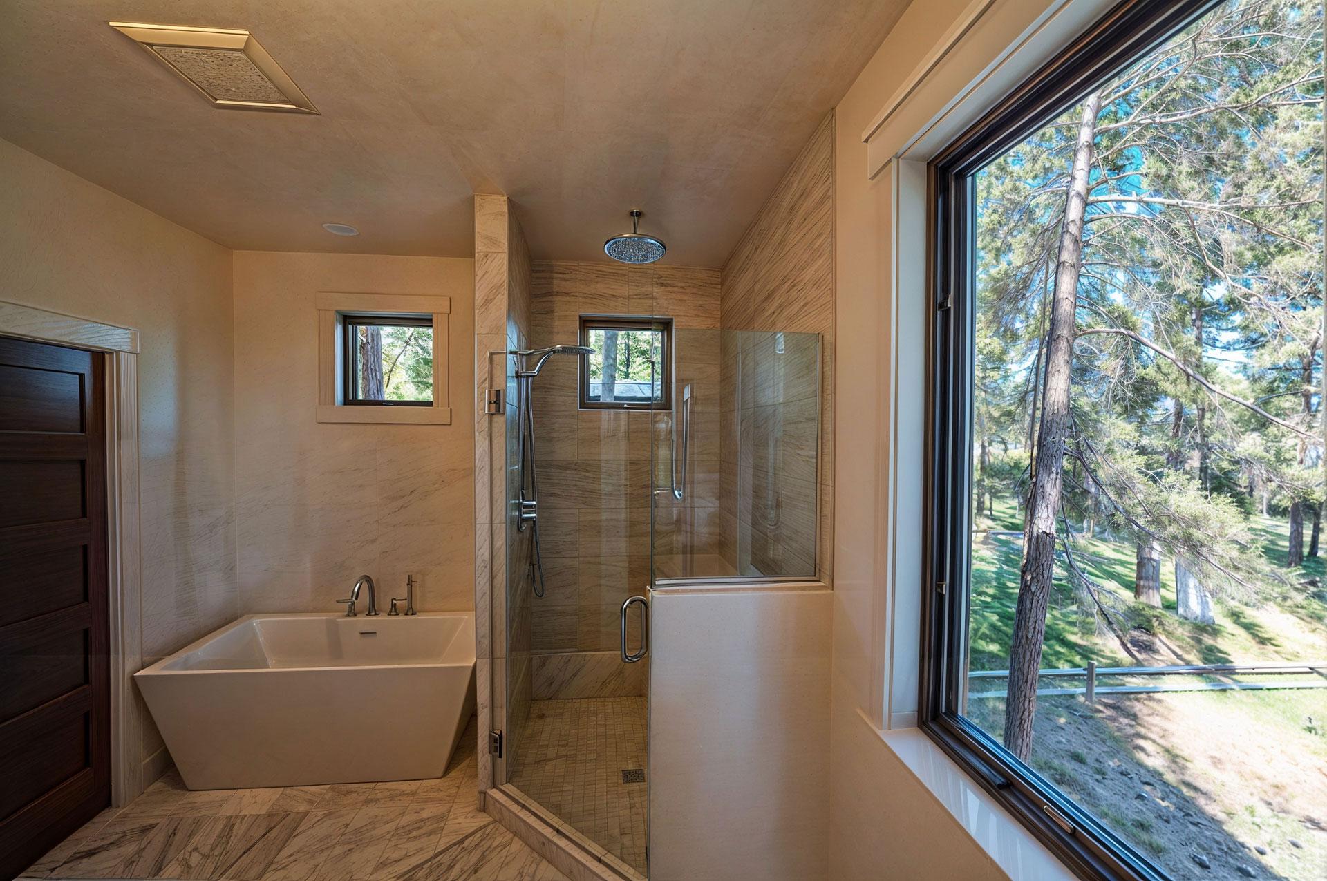Modern bathroom with freestanding tub, glass-enclosed shower with rain showerhead, and large window with forest view.