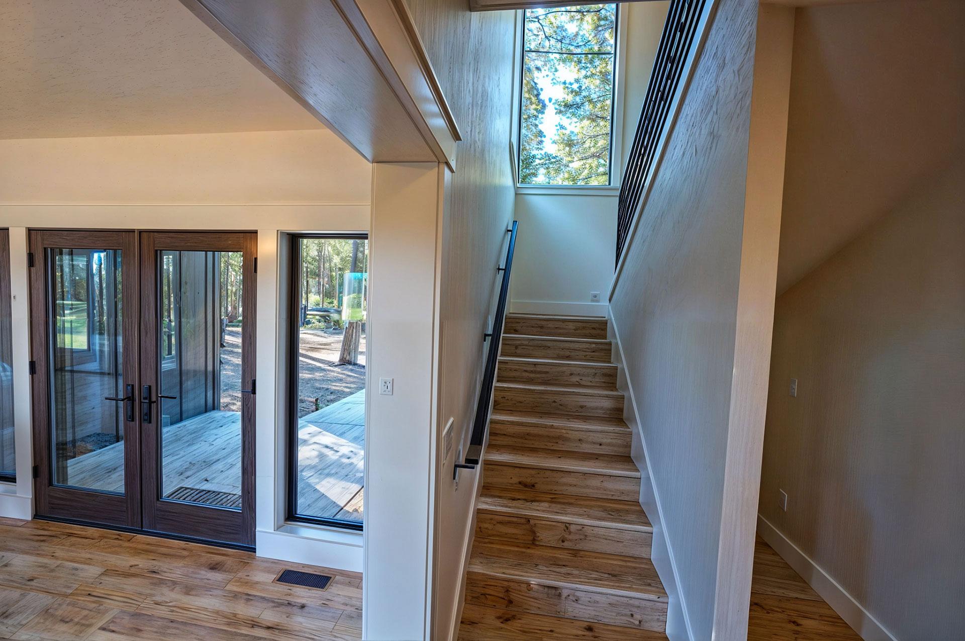 Interior view of wood plank staircase with large vertical window and double glass doors leading to exterior deck.