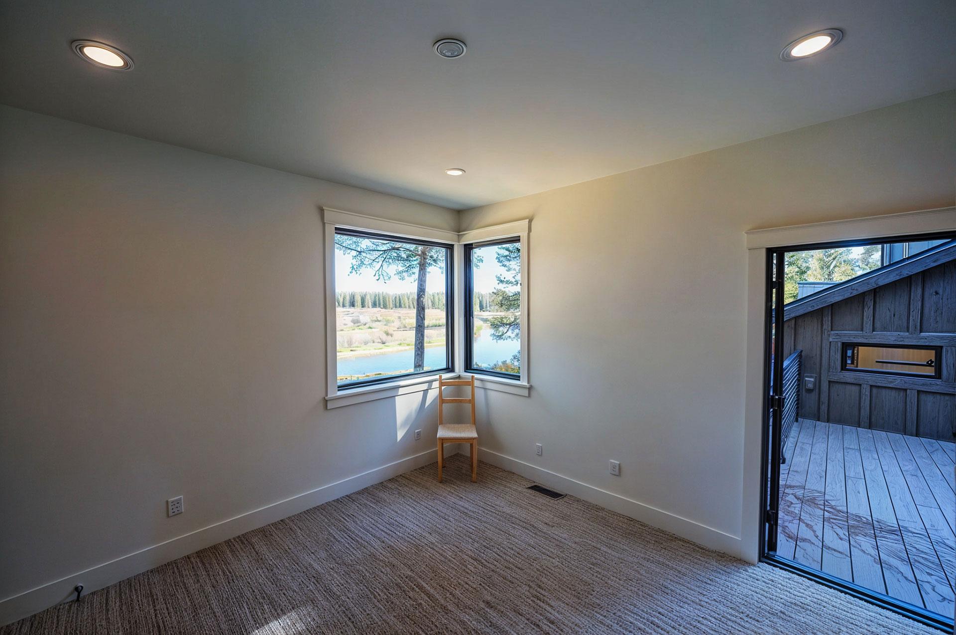 Interior room with corner windows overlooking a lake and trees. Doorway opens to a wooden deck.